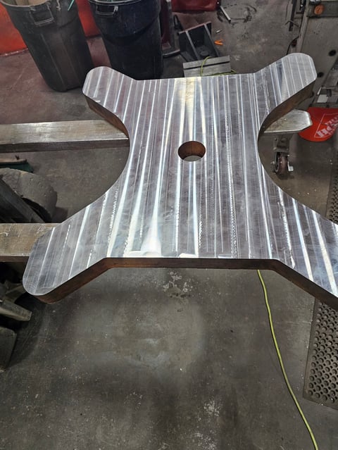 Metal cross-shaped bracket with striped pattern and center hole, positioned on a concrete workshop floor