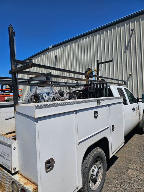 White pickup truck with service bed and roof rack loaded with coiled cables parked outside an industrial warehouse