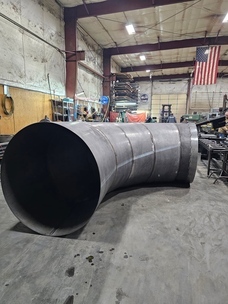 Large curved metal pipe in industrial warehouse with American flag visible in background