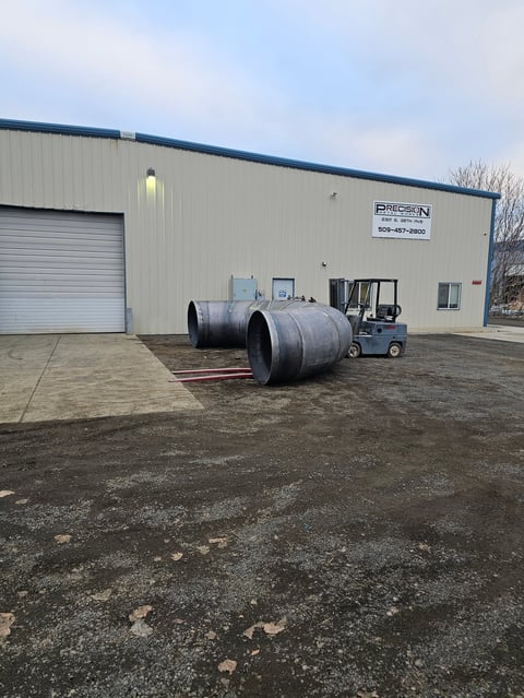 Forklift operator loading large cylindrical metal tank at industrial warehouse with white metal building