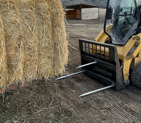 Skid steer loader with forks positioned to move a large hay bale on a farm yard